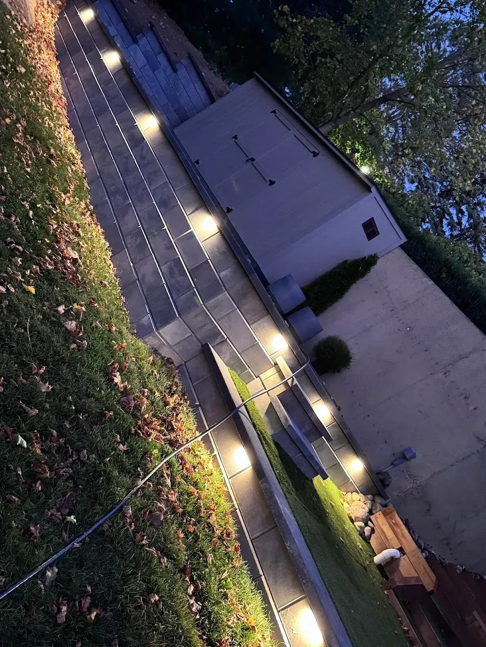 A paved garden path illuminated by small, round ground lights at dusk, leading toward a shed next to a grassy lawn.