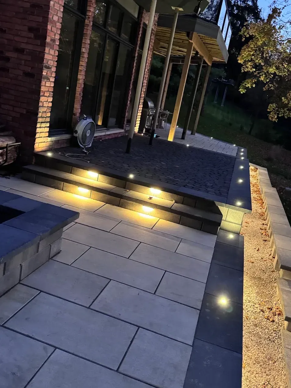 An outdoor patio featuring gray stone pavers, a raised dark-stone landing with built-in deck lights, and a brick house.