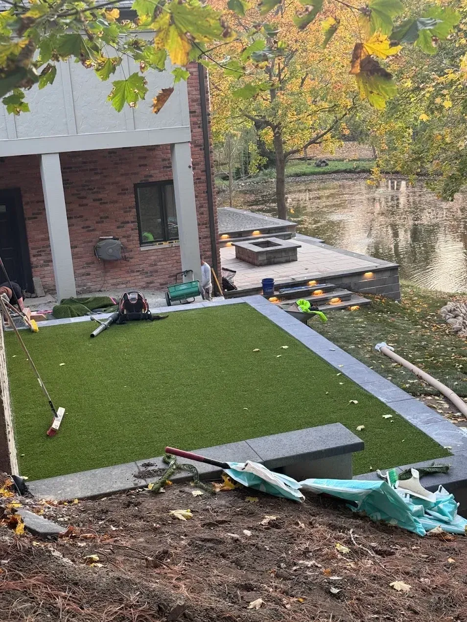 A newly installed patch of artificial turf bordered by gray stone sits next to a brick house with a fire pit by a pond.