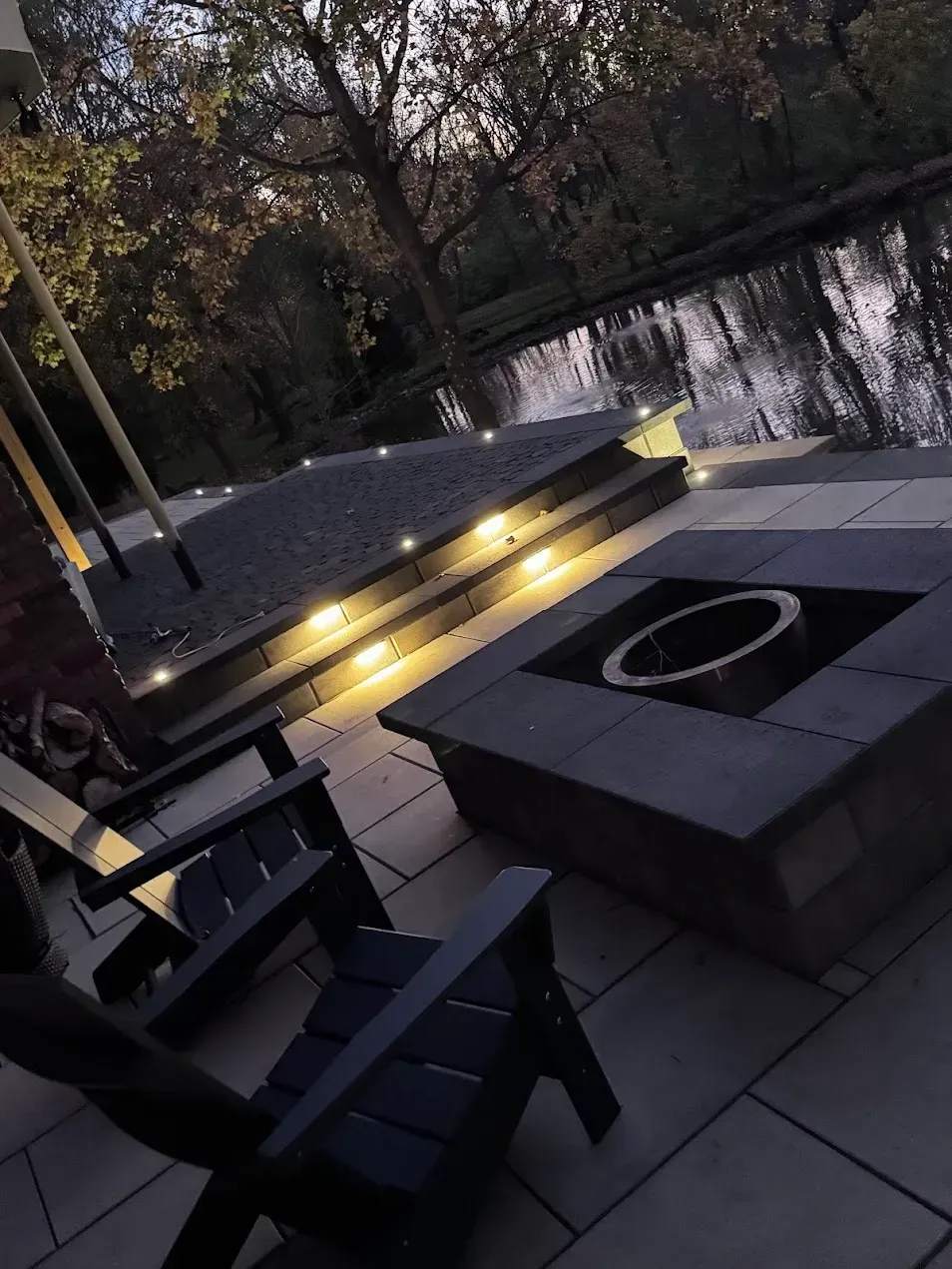 An outdoor patio at dusk featuring two dark chairs, a square fire pit, and illuminated stone steps overlooking a pond.