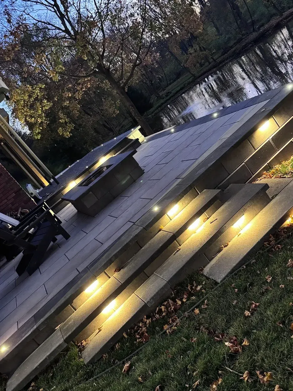 A multi-level stone patio at dusk with integrated LED stair lighting, overlooking a pond with a fire pit nearby.