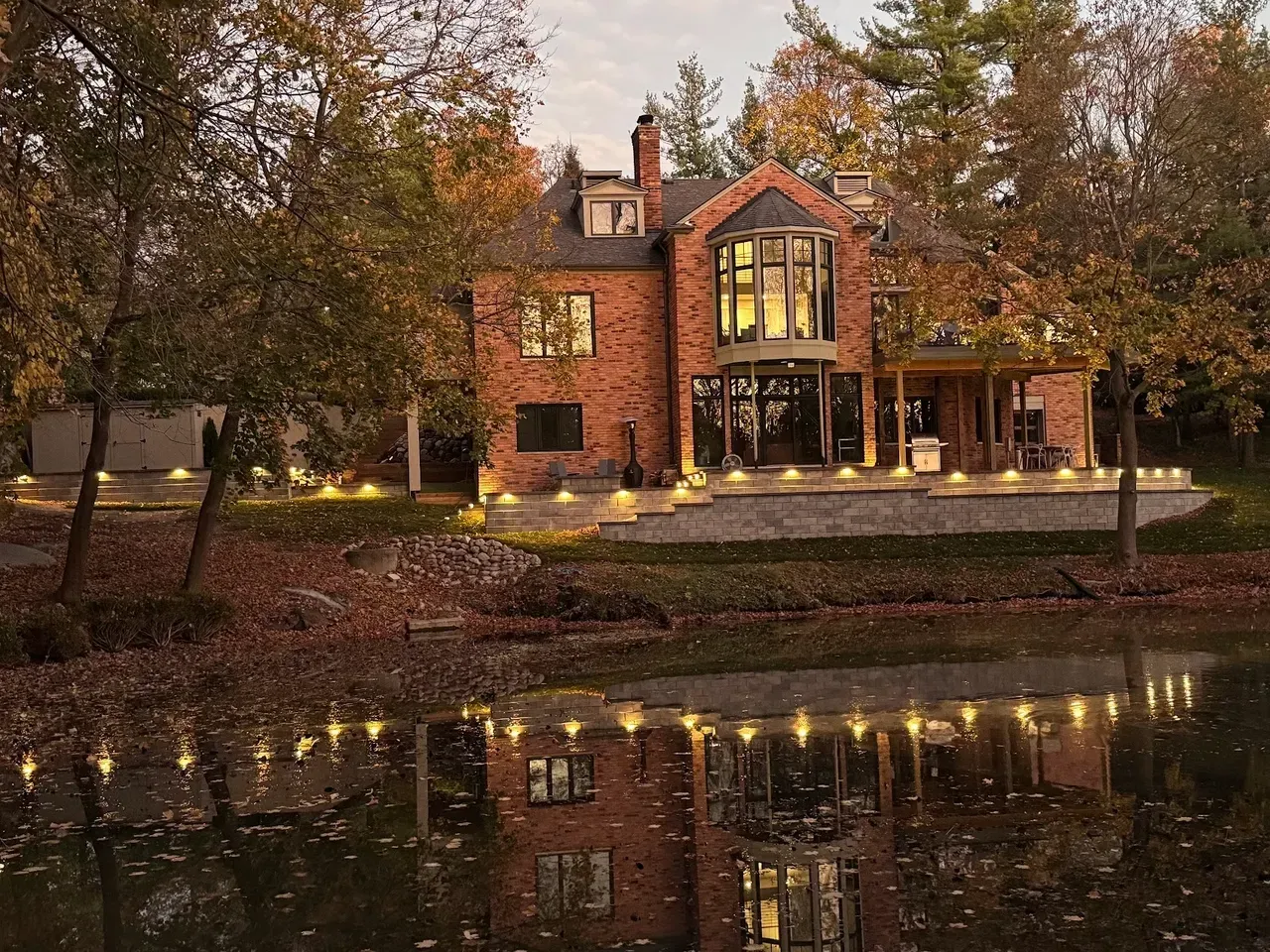 A brick house at dusk, illuminated by landscape lights and reflected in a calm pond, surrounded by autumn trees.