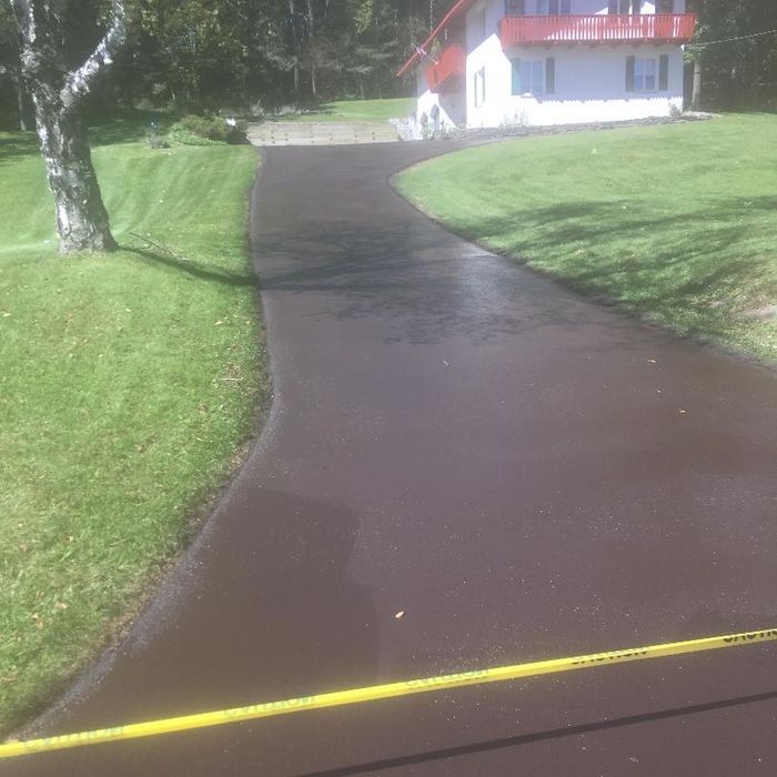 A freshly paved, dark asphalt driveway curves toward a house, marked off with a yellow caution tape in the foreground.