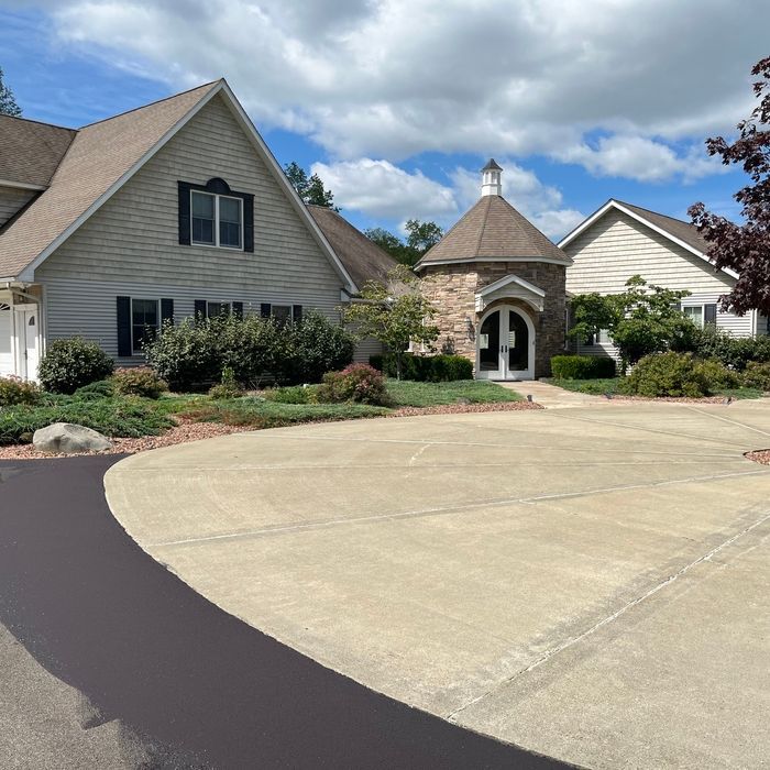 A suburban home with light siding, a stone turret entry, a large circular concrete driveway, and lush green landscaping.