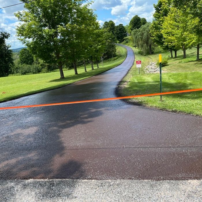 A freshly paved, dark asphalt driveway winds uphill through a lush green landscape with trees, marked by orange tape.