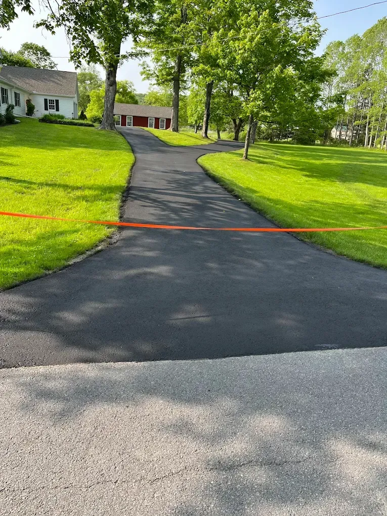 A freshly paved black asphalt driveway leads up a grassy yard toward a white house with an orange caution tape across it.
