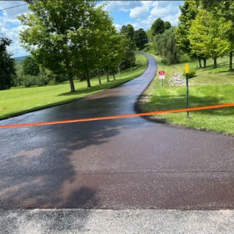 A freshly paved, dark asphalt driveway curves uphill through a grassy lawn with trees, blocked by an orange tape barrier.