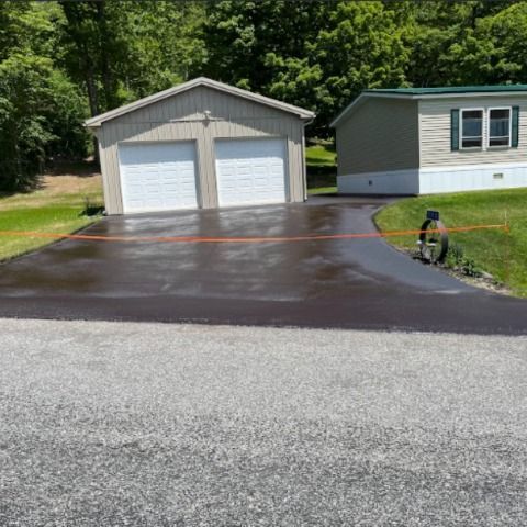 A freshly sealed dark asphalt driveway leads to a two-car garage and house, with a bright orange caution tape across it.