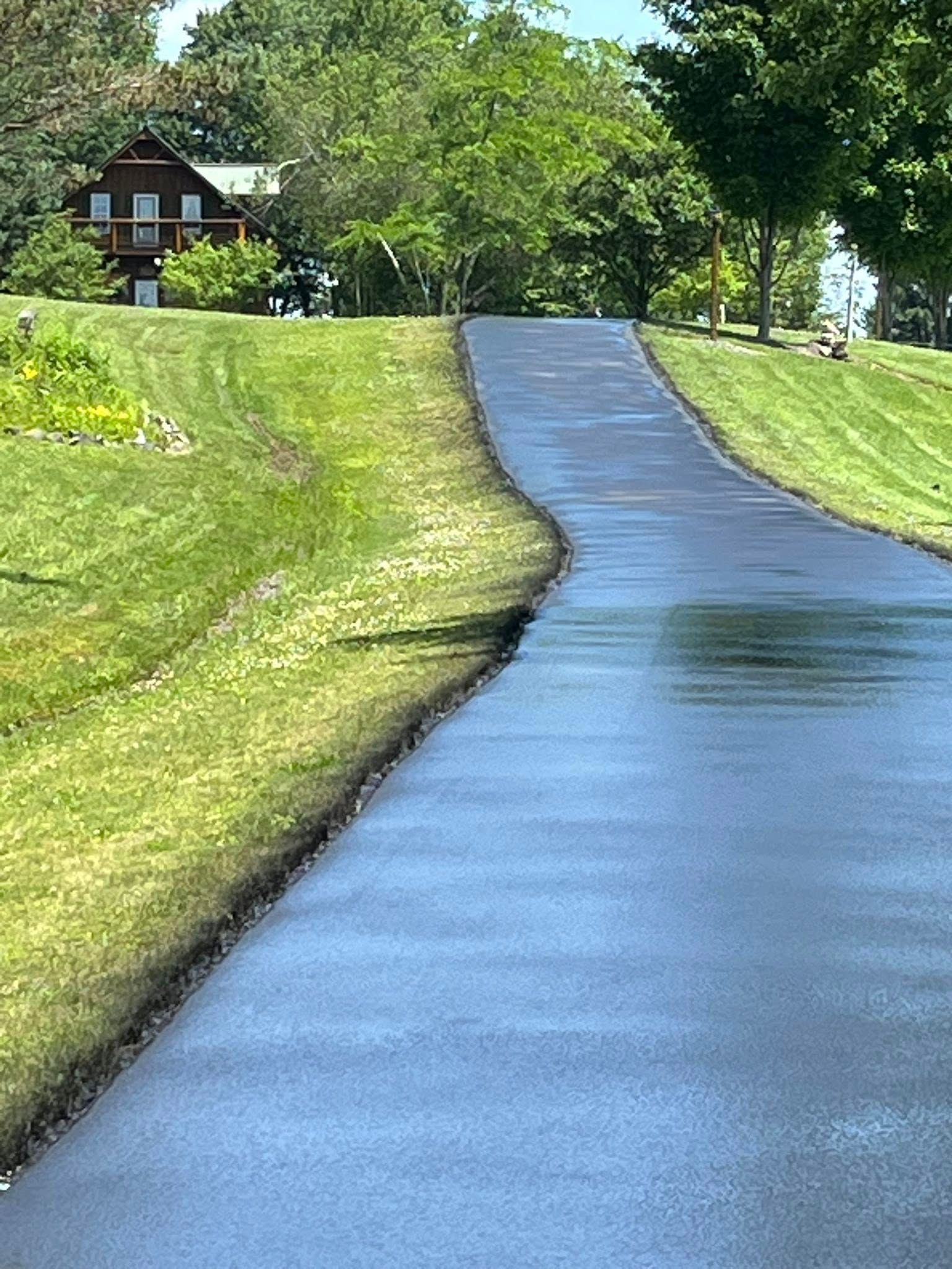 A freshly paved, dark asphalt driveway leads uphill through a grassy lawn toward a wooden house nestled in green trees.