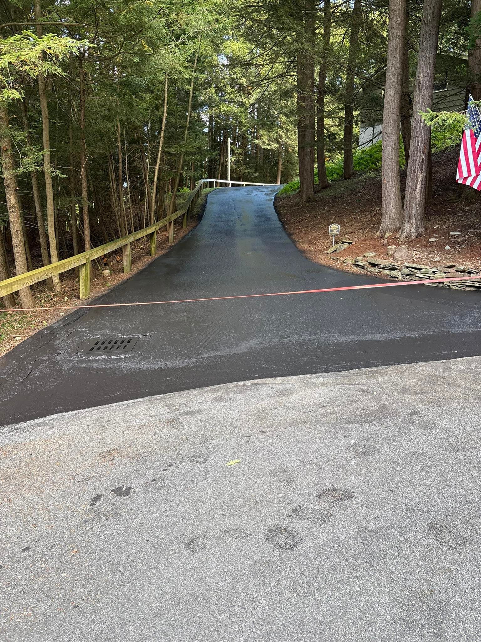 A freshly paved asphalt driveway transitions from a gravel area into a wooded property, marked by a painted white line.