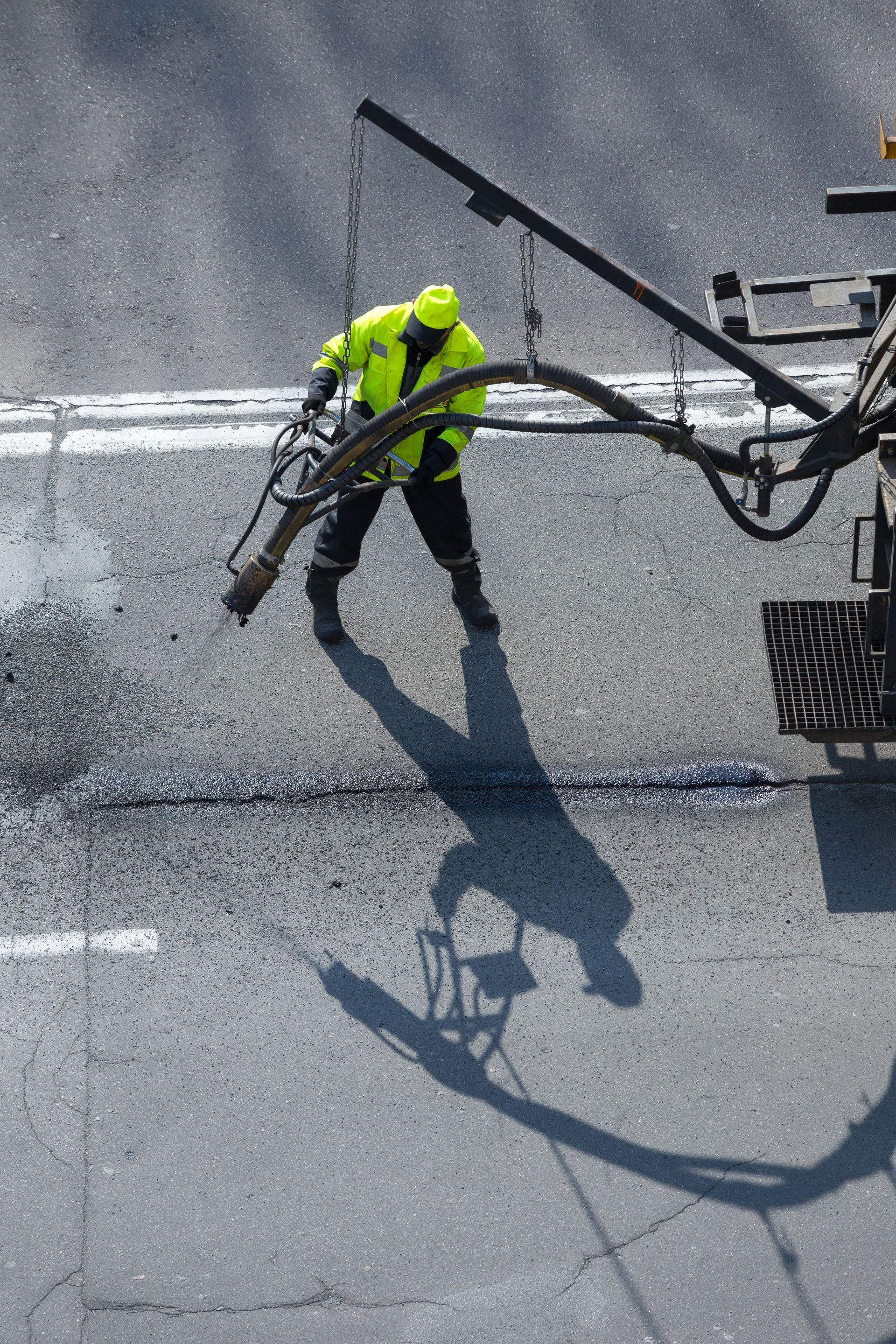 A worker in a bright yellow jacket uses a truck-mounted spray hose to repair a crack in an asphalt road.