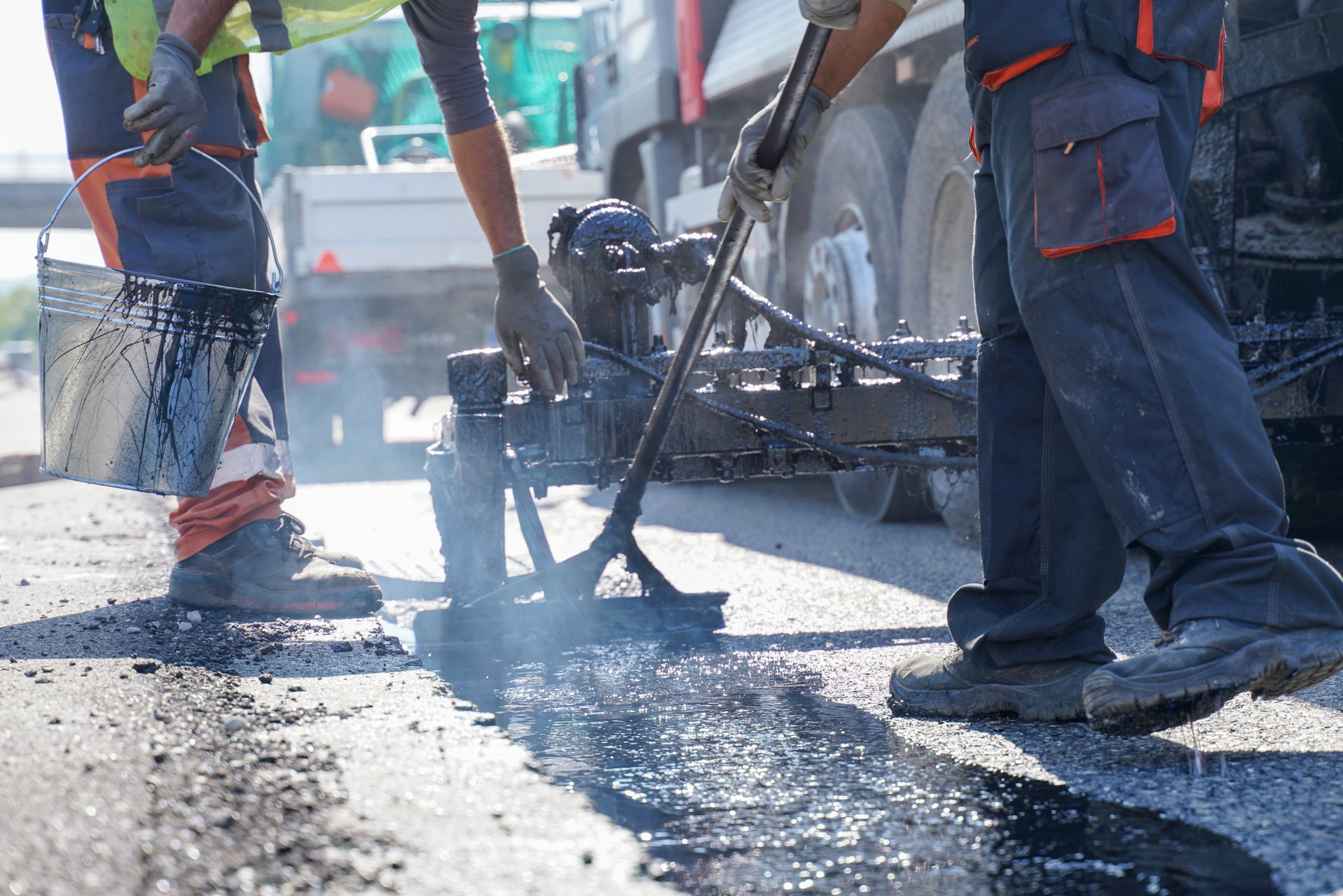 Two workers in high-visibility clothing repairing a road with hot asphalt and specialized tools.
