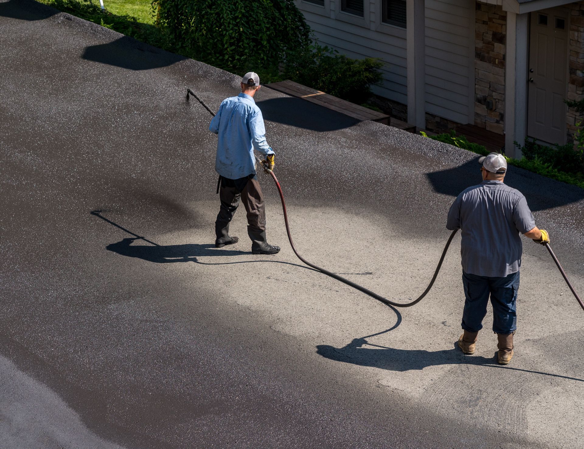 Two workers use a hose to apply sealant to a driveway in front of a house.