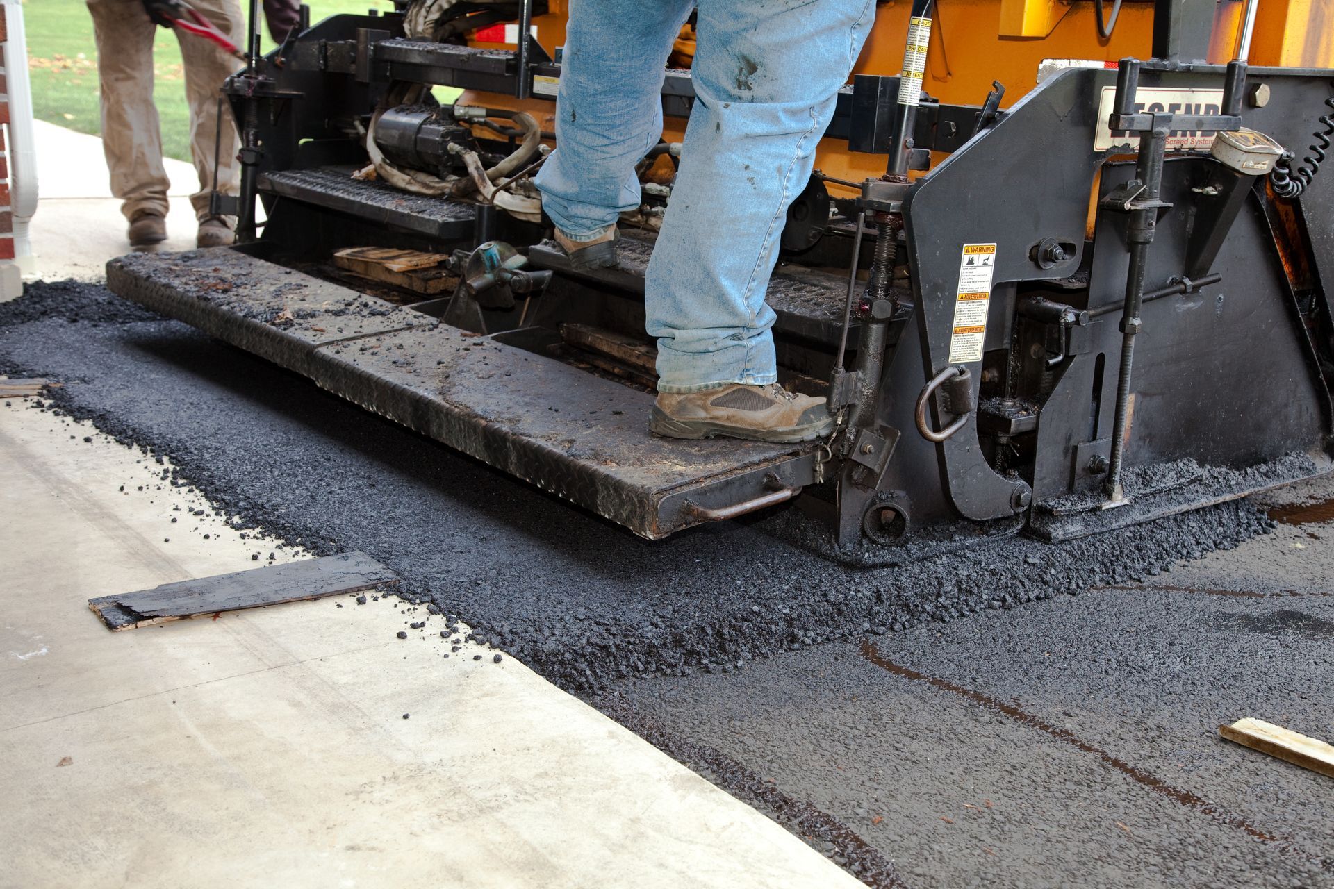 A construction worker stands on a paving machine as it lays fresh black asphalt on a surface next to a concrete walkway.