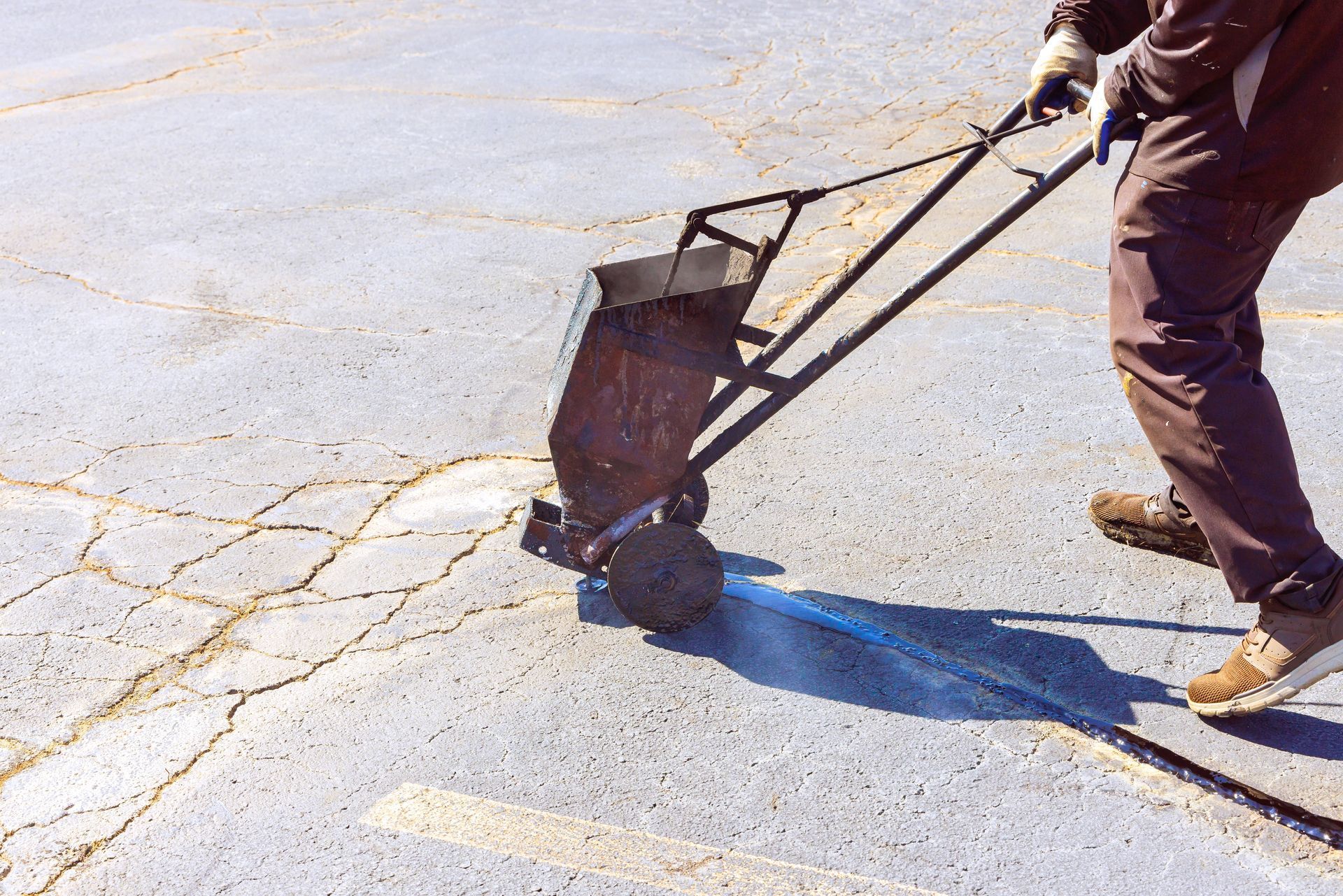 A worker uses a rolling machine to apply sealant to cracks on an asphalt parking lot surface.