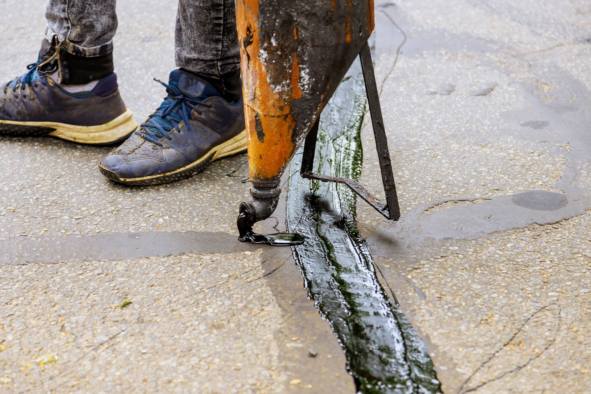 A person uses a crack sealing machine to pour hot, liquid asphalt into a crack on a concrete road surface.