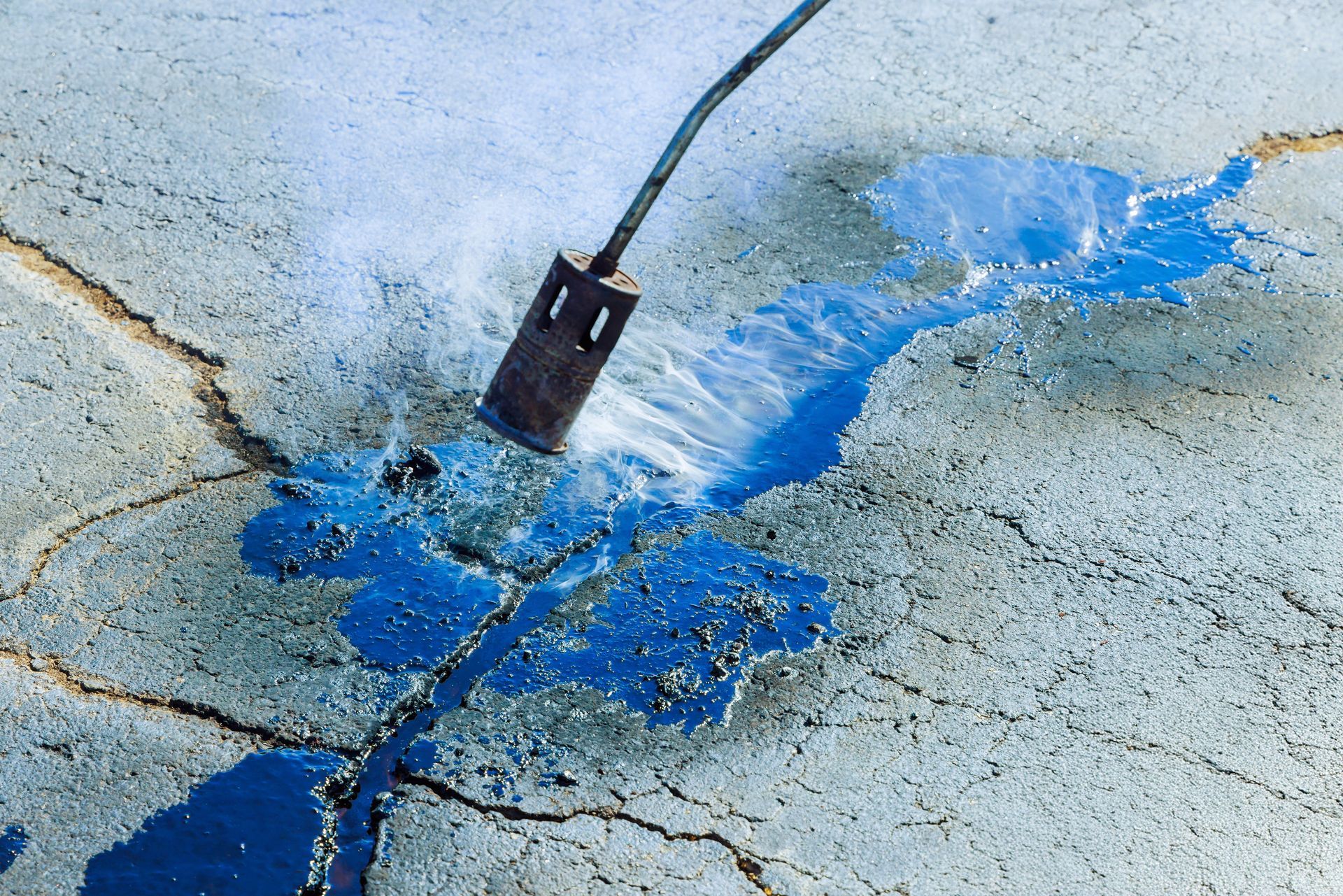 A propane torch heats a bright blue, liquid material filling deep cracks in a grey concrete surface.