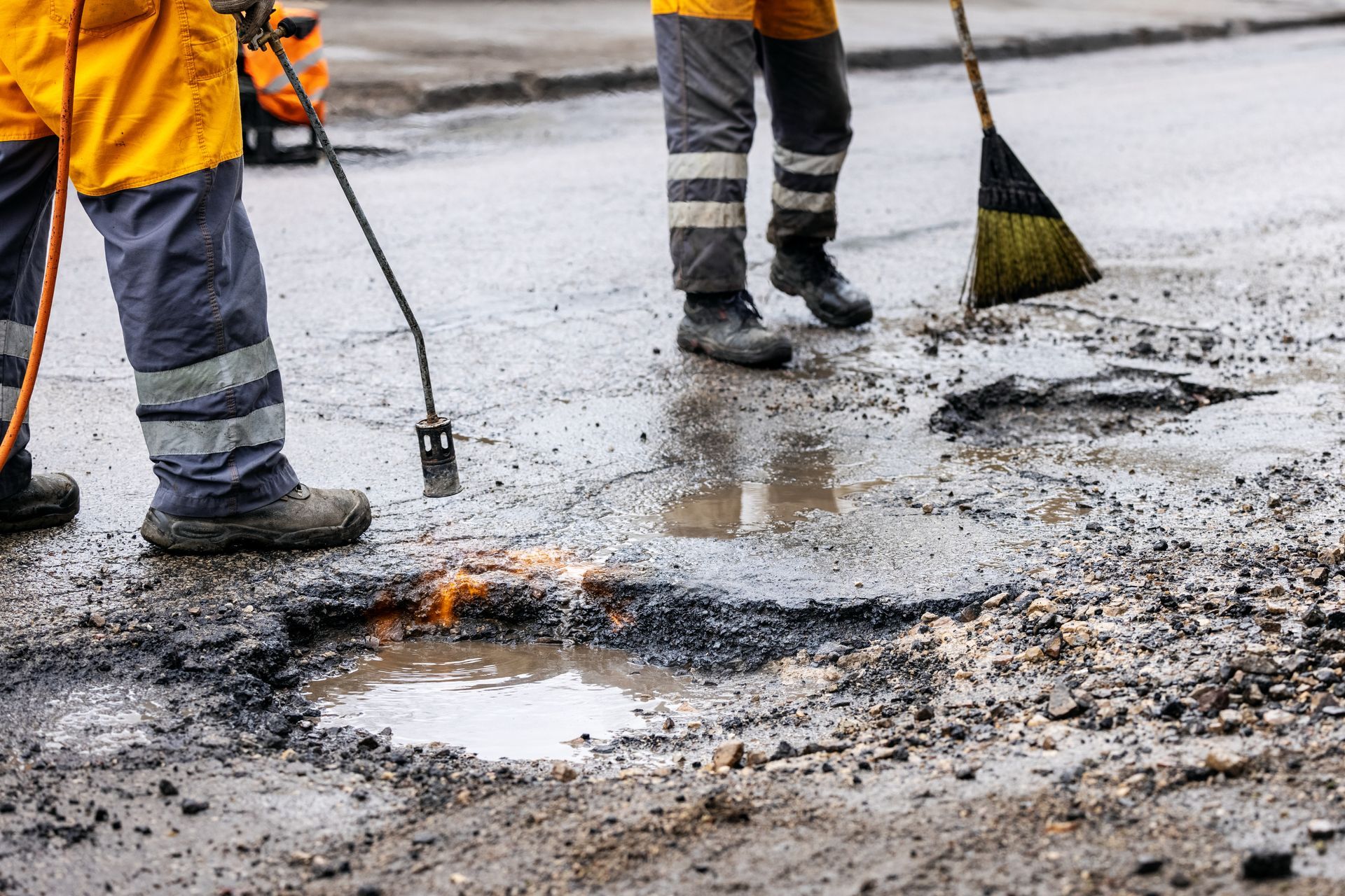 Workers in high-visibility orange clothing use a heat torch and a broom to repair a pothole in a wet, damaged road.