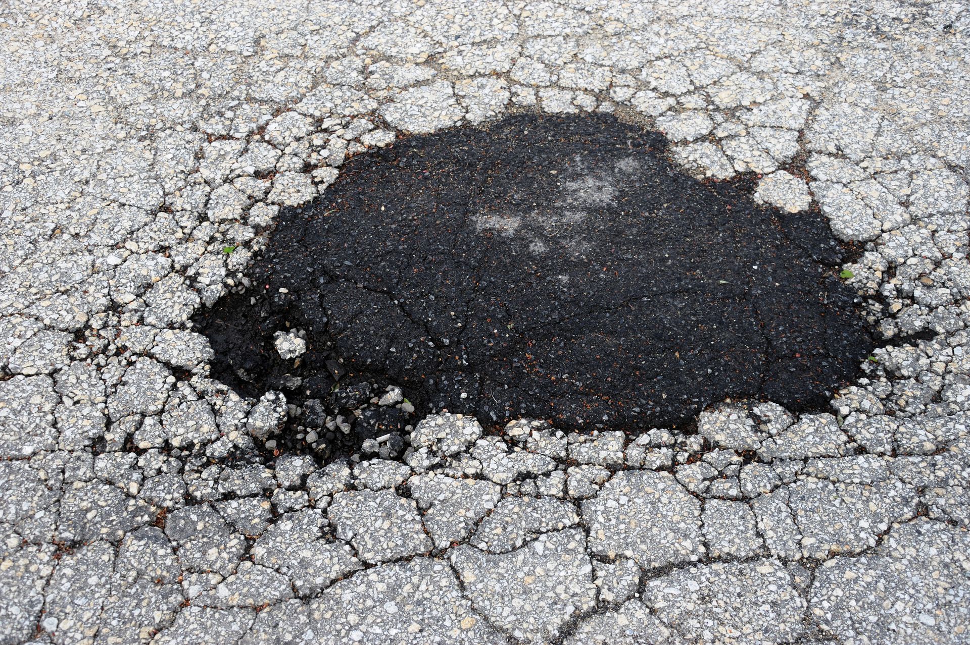 A large, dark pothole surrounded by a web of deep cracks in grey asphalt pavement.