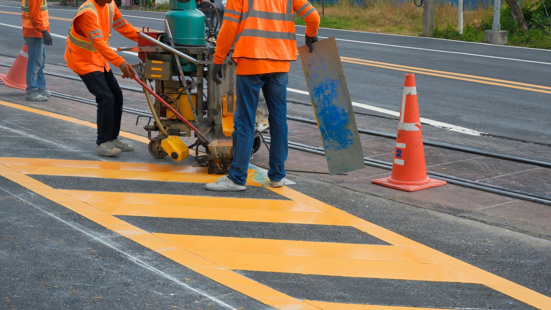 Two workers in high-visibility orange vests use machinery to paint yellow road markings on asphalt near a traffic cone.