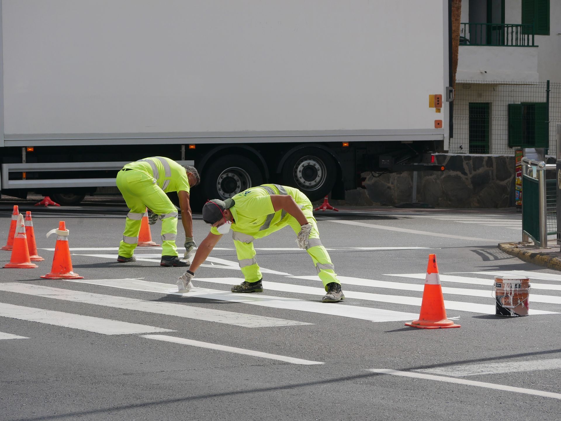 Two workers in neon yellow uniforms paint white stripes on an asphalt crosswalk, surrounded by orange traffic cones.