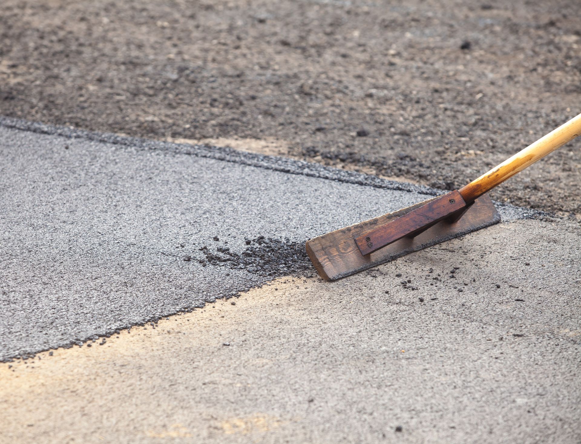 A long-handled asphalt lute spreading fresh, dark gravel over a lighter-colored pavement surface.