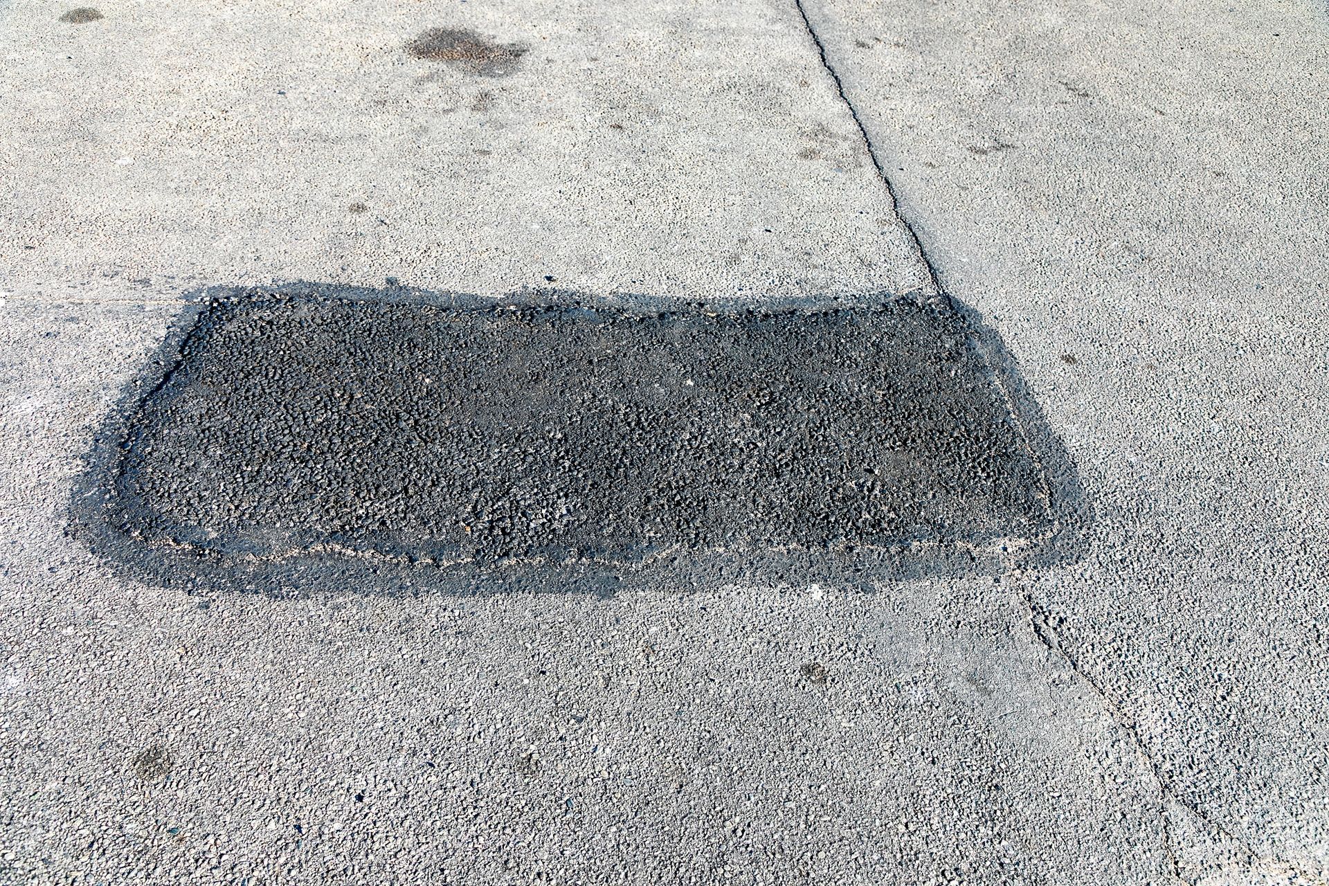 A rectangular patch of dark asphalt filling a hole in a light-colored concrete sidewalk.