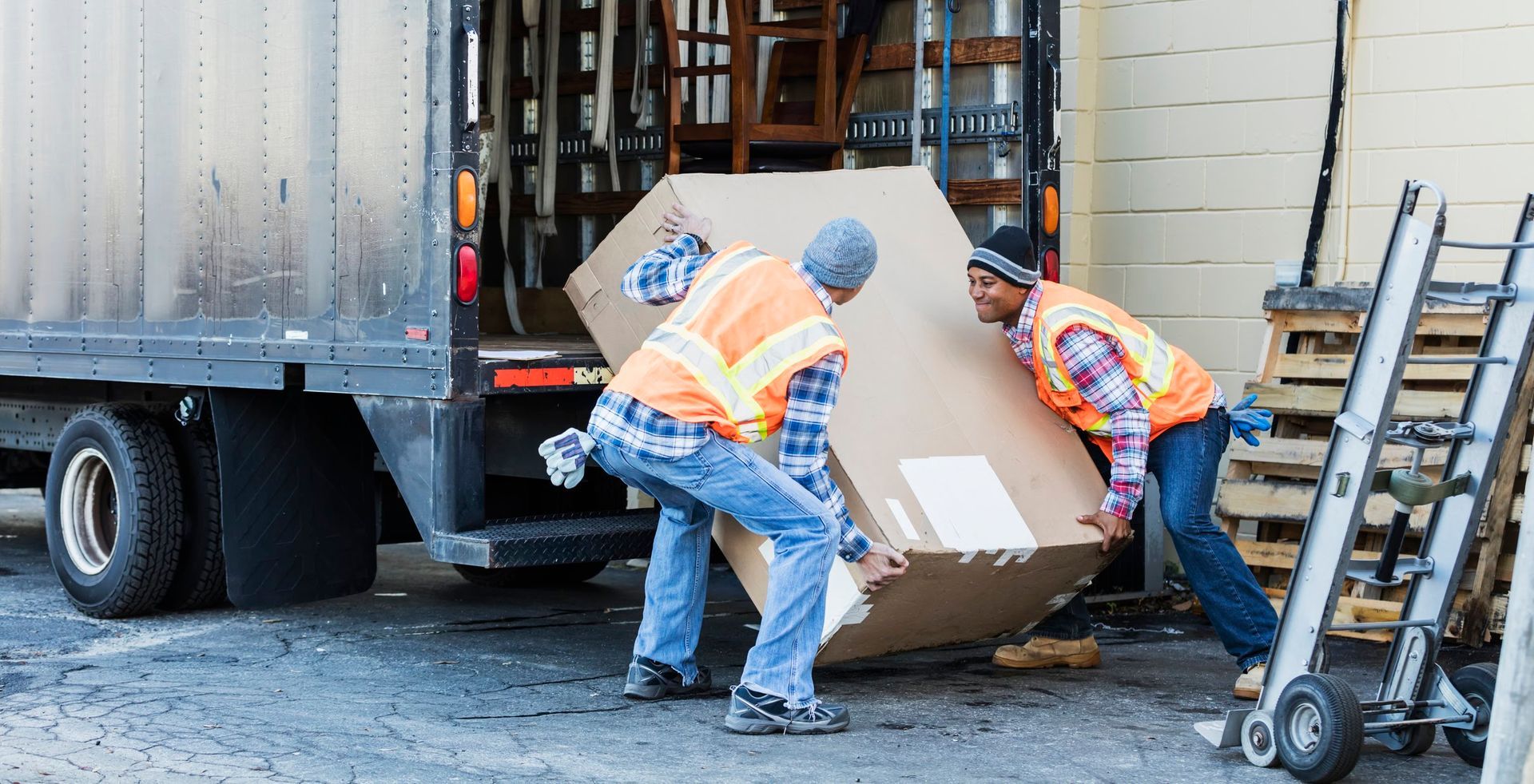 Due operai con giubbotti di sicurezza caricano una grande scatola di cartone da un camion.