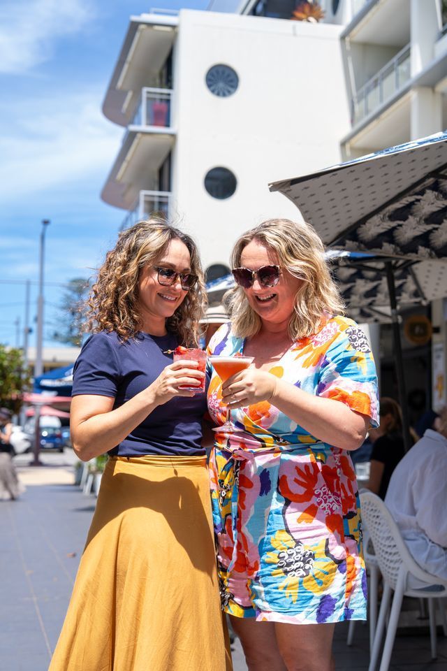 Two women clink drinks on a sunny day. Building and outdoor cafe in the background.