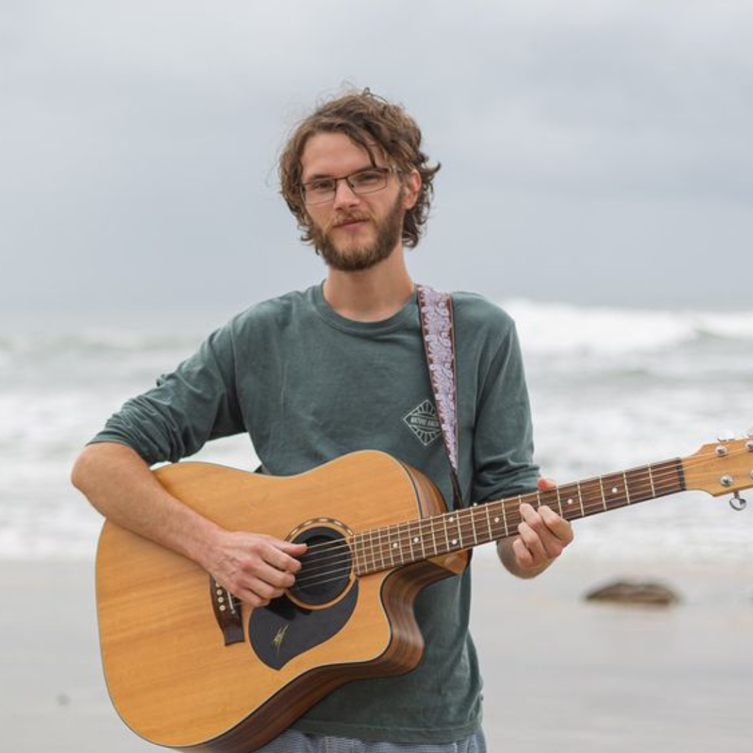 A Man Is Holding an Acoustic Guitar on The Beach — Chop 'n Chill in Port Macquarie, NSW