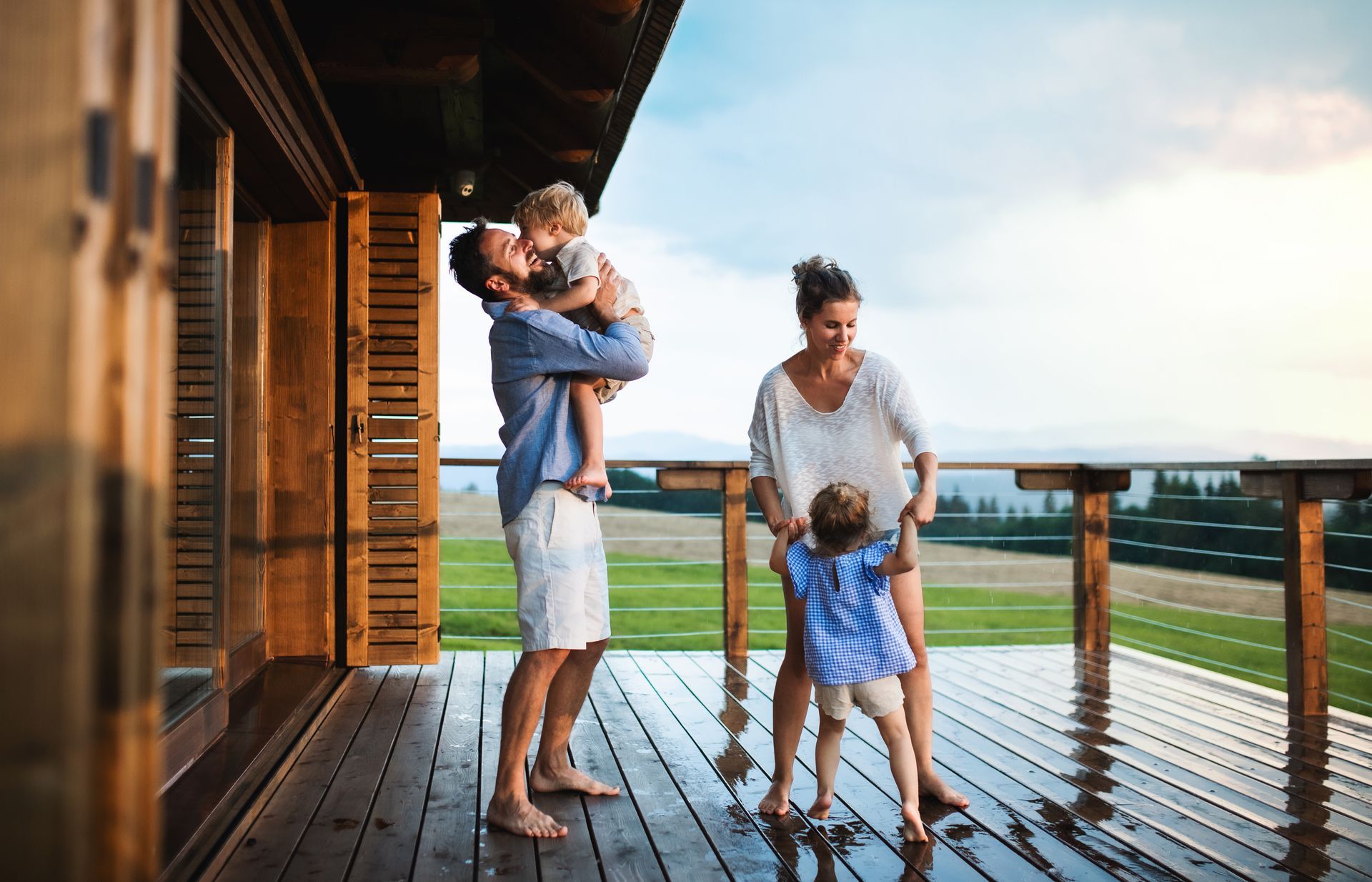 Family playing on a wooden deck with a scenic view: man holding child, woman with another child.
