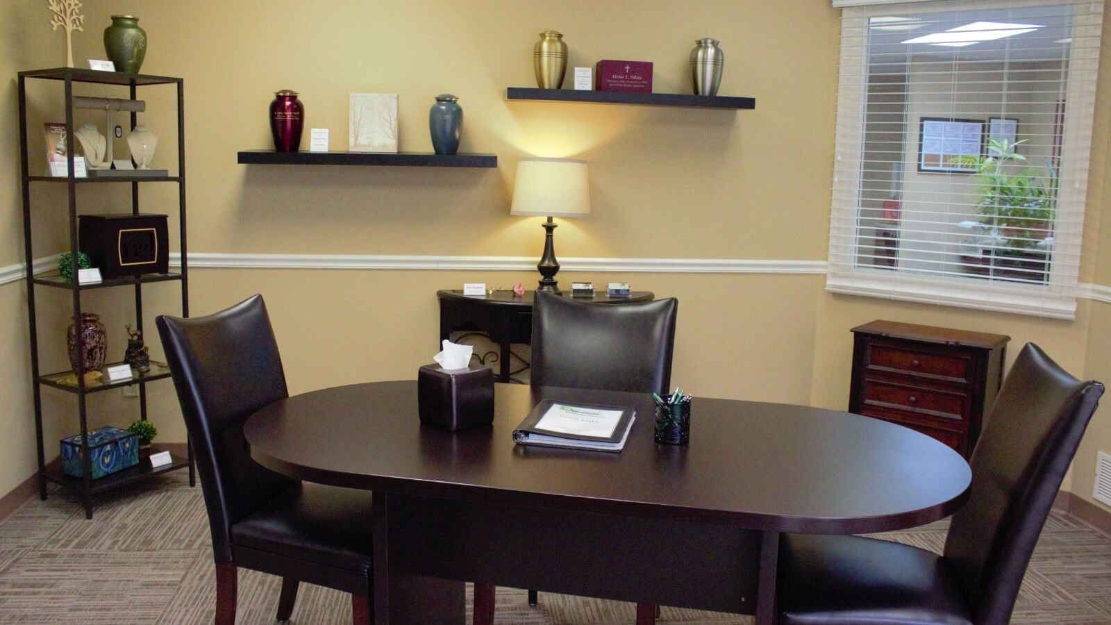 Small office conference room with oval table, chairs, shelves, lamp, and window blinds