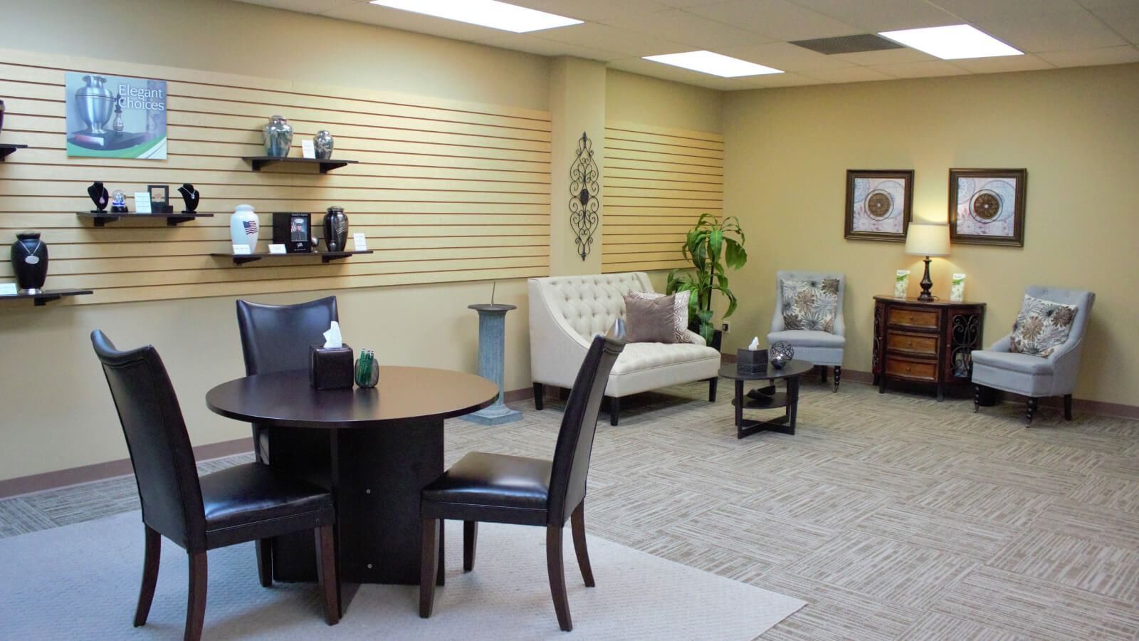 Small waiting room with round table, chairs, beige walls, display shelves, and framed art.