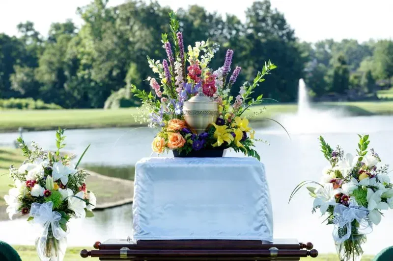 Floral arrangement on a white-draped table by a lake with a fountain and trees in the background