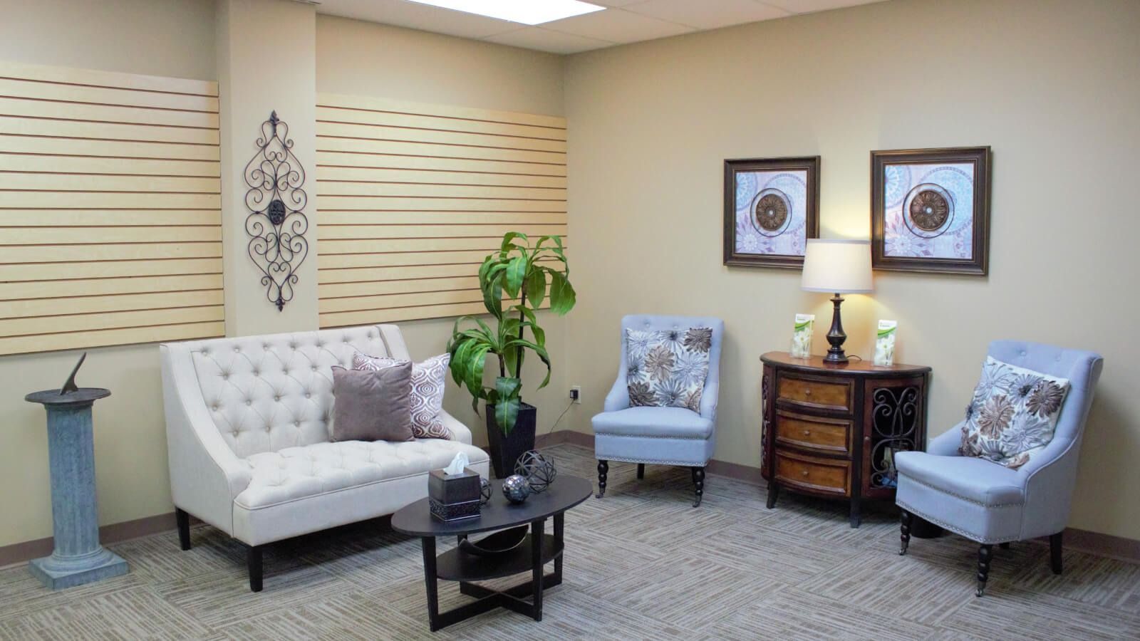Comfortable waiting room with white and blue chairs, small table, plant, and framed art on beige walls.