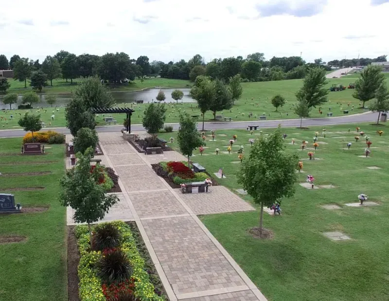Landscaped cemetery with a stone walkway, flower beds, and rows of headstones under a cloudy sky