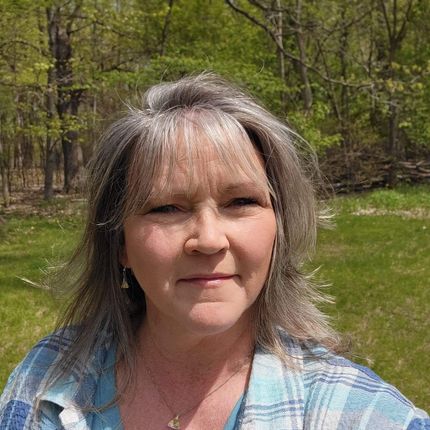 A woman with gray hair is standing in a field with trees in the background.