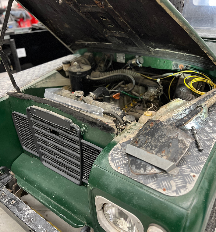 An open hood of a green Land Rover, revealing the engine bay, a black radiator grill, and a shovel on the fender. | Kensington Motor Cars