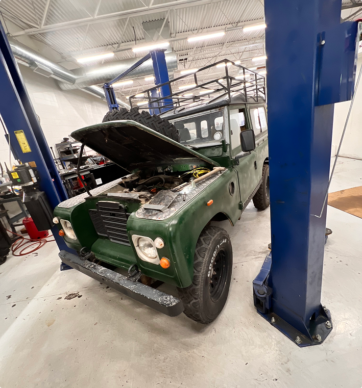 A dark green Land Rover with its hood open, parked inside an auto repair shop between two blue vehicle lift pillars. | Kensington Motor Cars