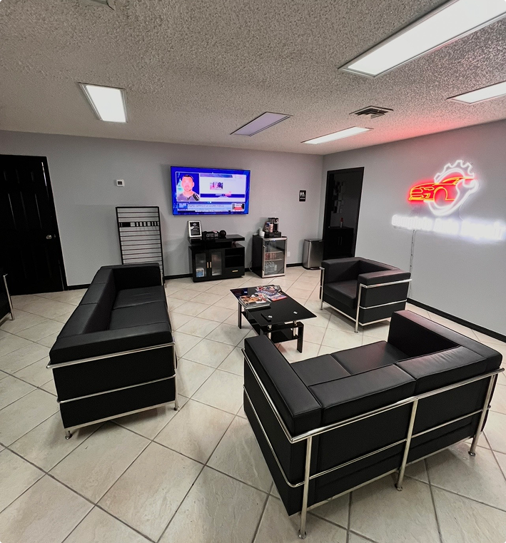 Modern office waiting room with black leather sofas, a coffee table, a wall-mounted TV, and a neon logo on the wall. | Kensington Motor Cars