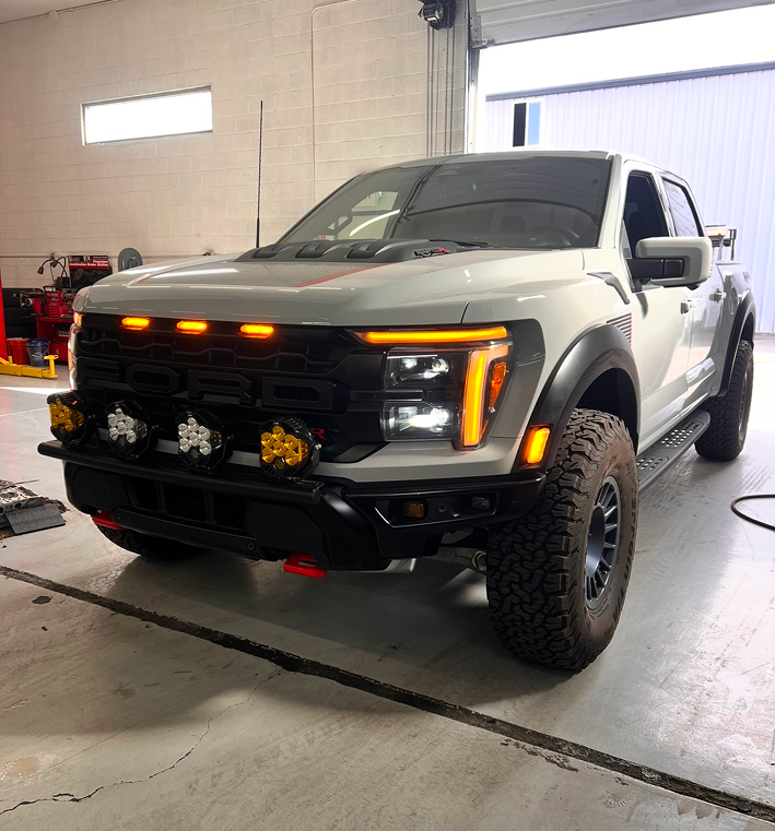 A white Ford F-150 Raptor pickup truck with aftermarket off-road lights parked inside an auto shop garage. | Kensington 