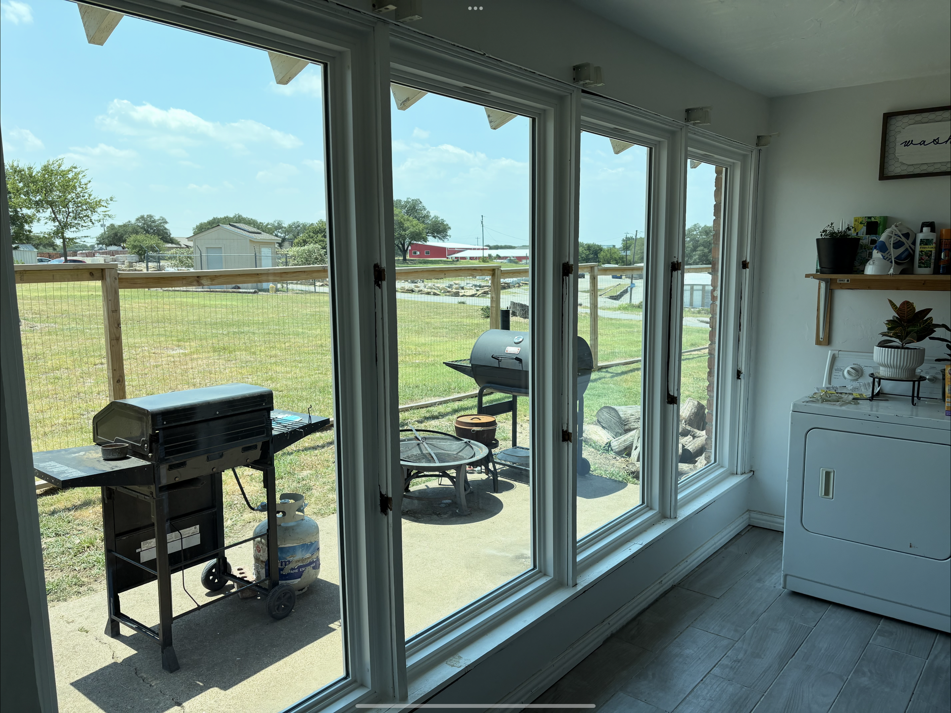 View from inside looking out large windows at a backyard with a grill, fence, and cloudy sky.