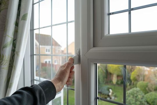 Hand opening a white window with grid panes, showing a garden and a house.