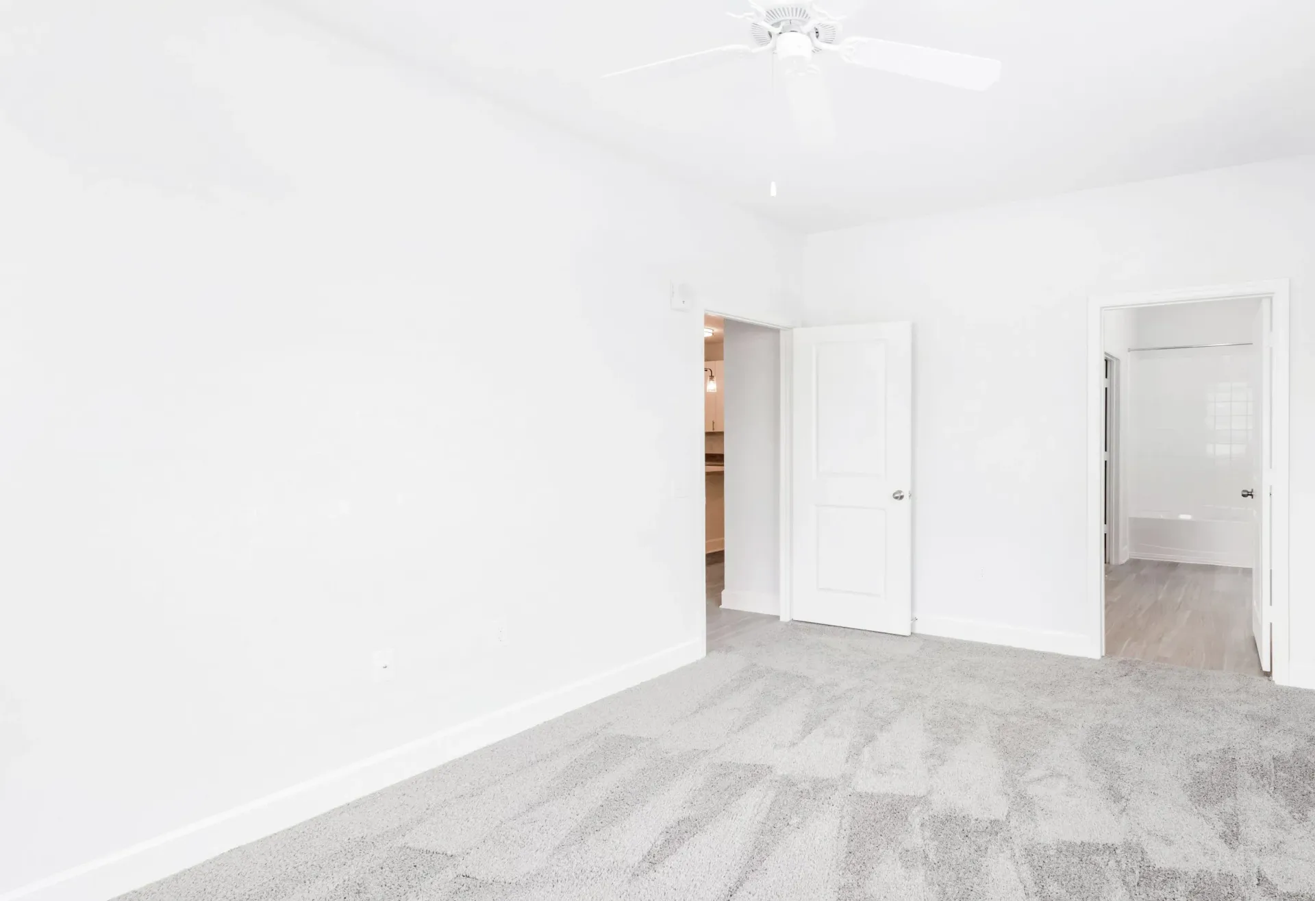 Empty bedroom with white walls, a ceiling fan, and light grey carpet; doors lead to adjacent rooms.