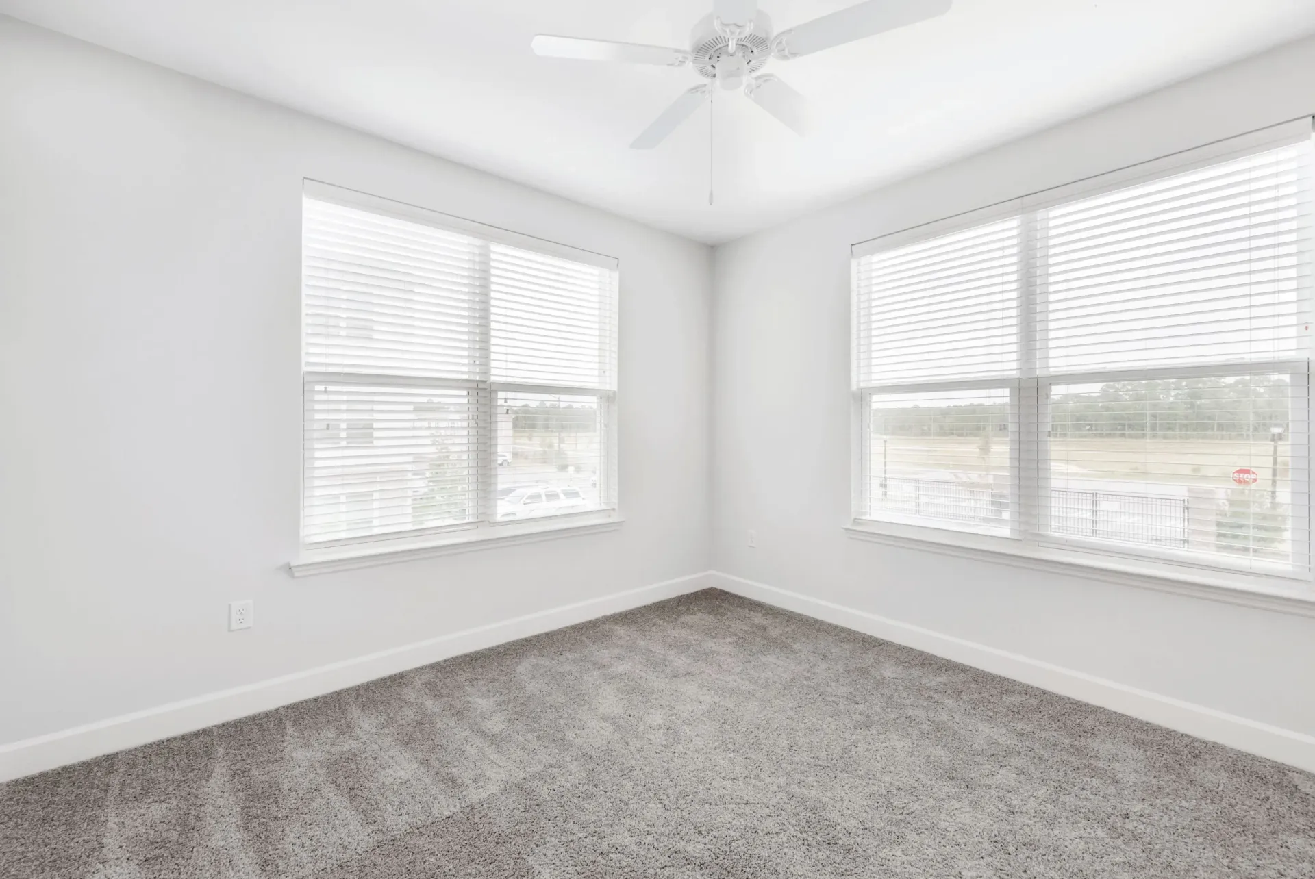 Bright empty bedroom with white walls, two large windows with blinds, and a ceiling fan.