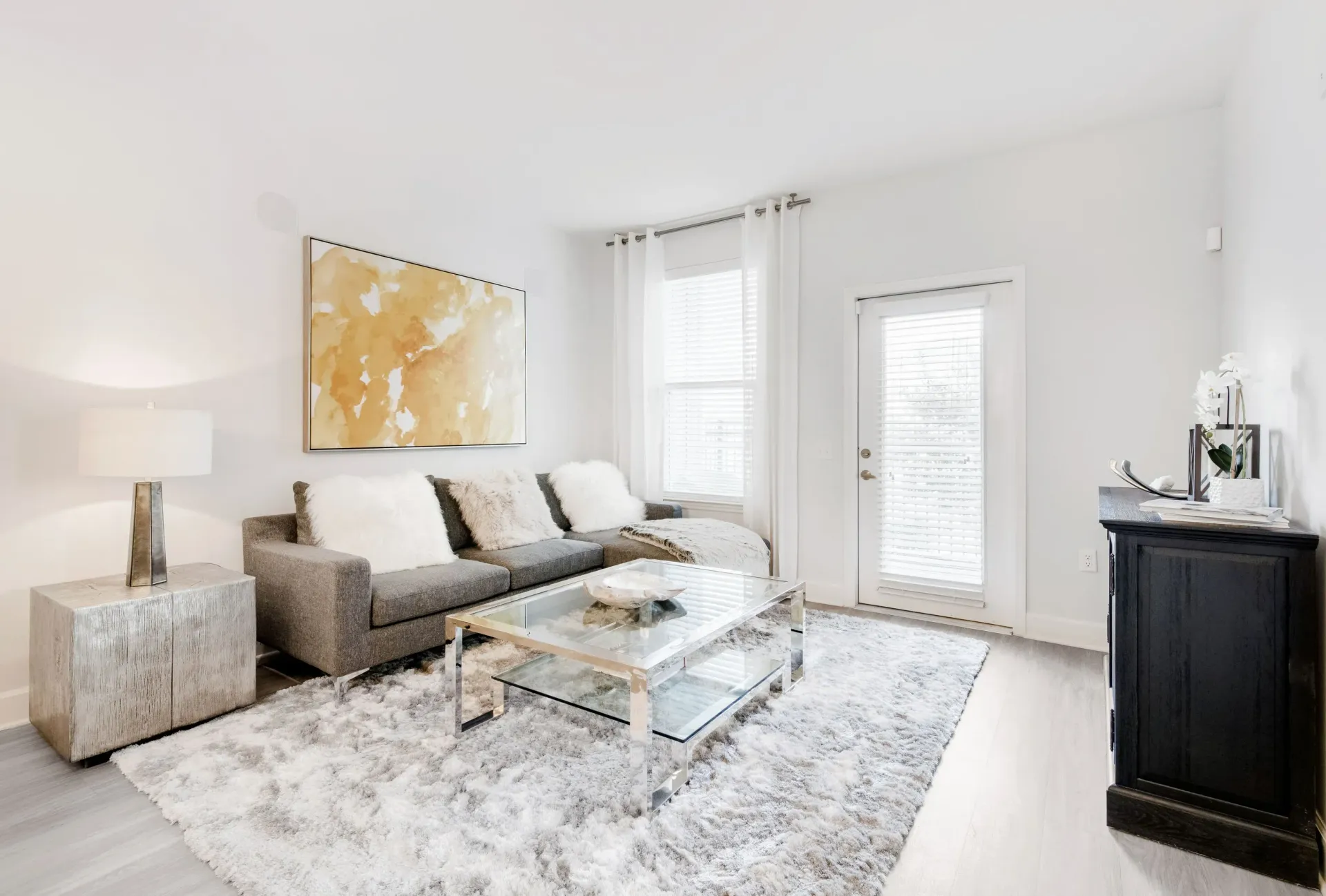 Bright living room with a gray sofa, glass coffee table, white curtains, and a plush rug.