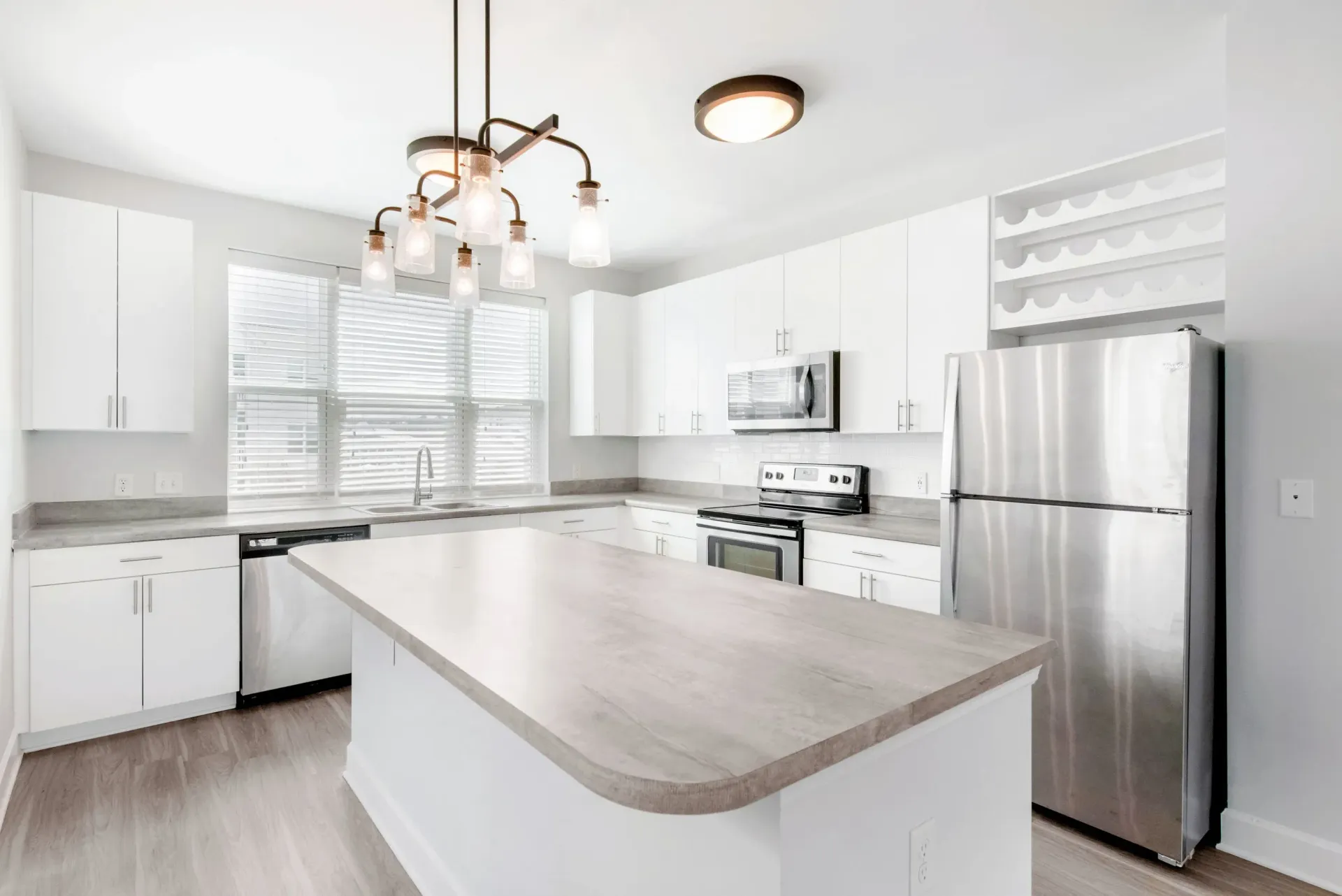 Bright white kitchen with island, stainless steel fridge/oven, and large window over the sink.