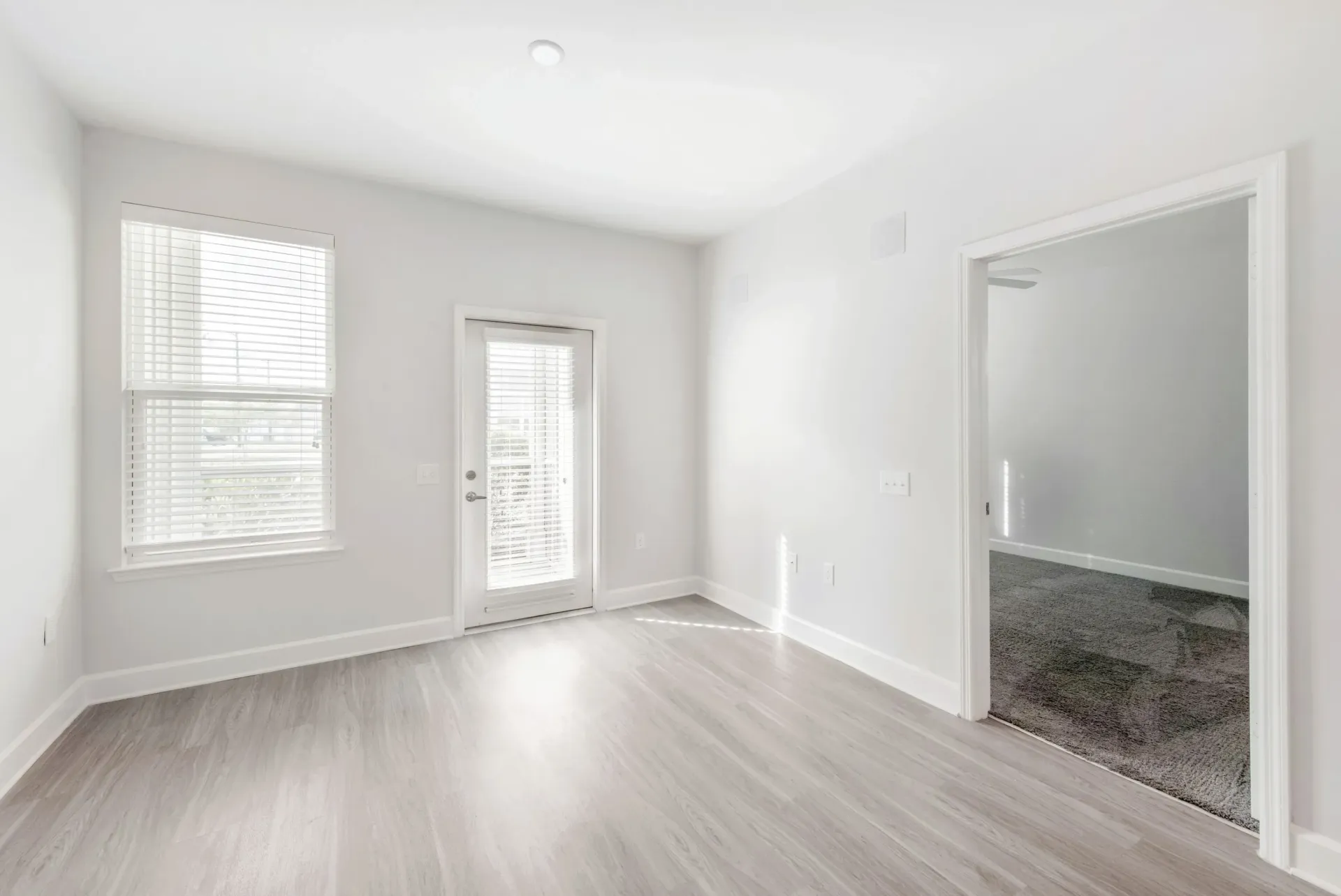 Empty apartment room with light wood floors, white walls, two windows with blinds, and a door to the outside.