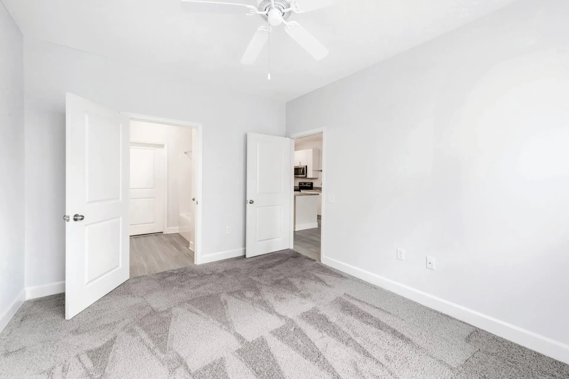 Empty bedroom with light gray walls, carpet, and a ceiling fan; doors open to a hallway and kitchen.