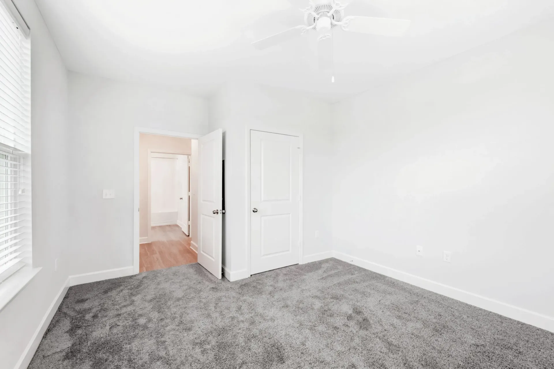 Empty bedroom with white walls, gray carpet, ceiling fan, window with blinds, and two doors.